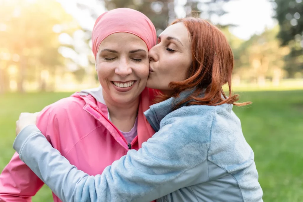 Two women hug outdoors; one in a pink headscarf smiles as she's kissed, with ReCenter hospital in the background.