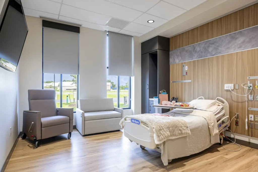 A welcoming hospital room with a bed, loveseat, TV, and wood-inspired décor provides a comfortable, concierge feel.