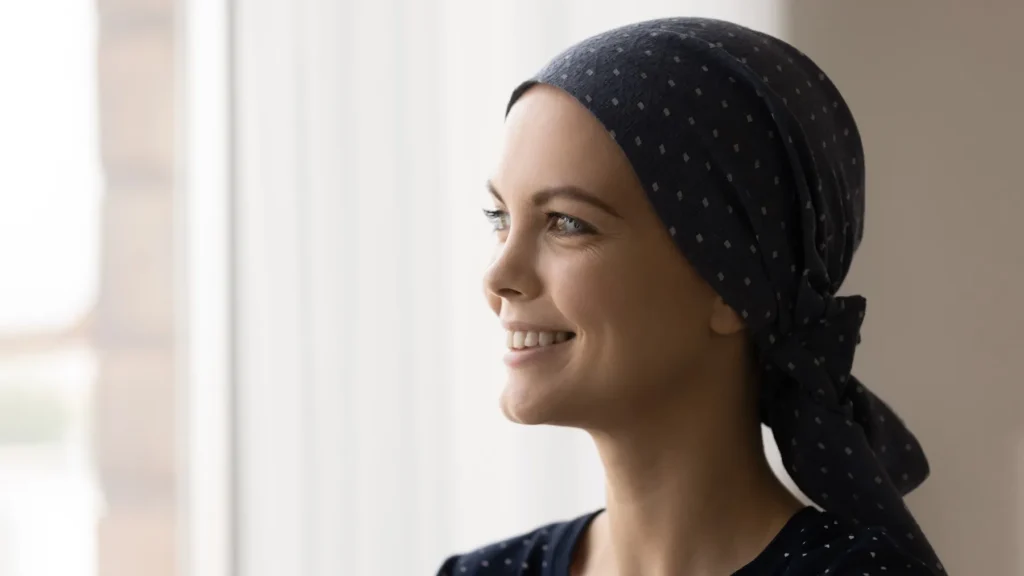 A woman wearing a dark headscarf with white dots smiles brightly by a window, expressing hope during her treatment.