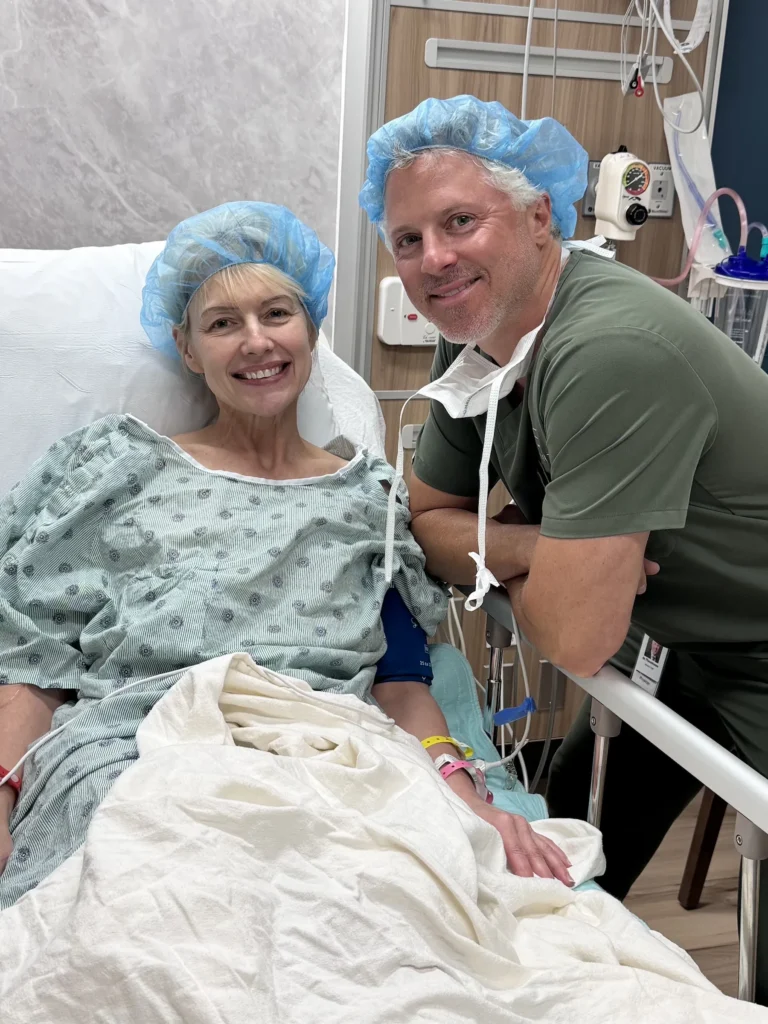 A woman in a hospital bed smiles beside a doctor, both wearing blue surgical caps