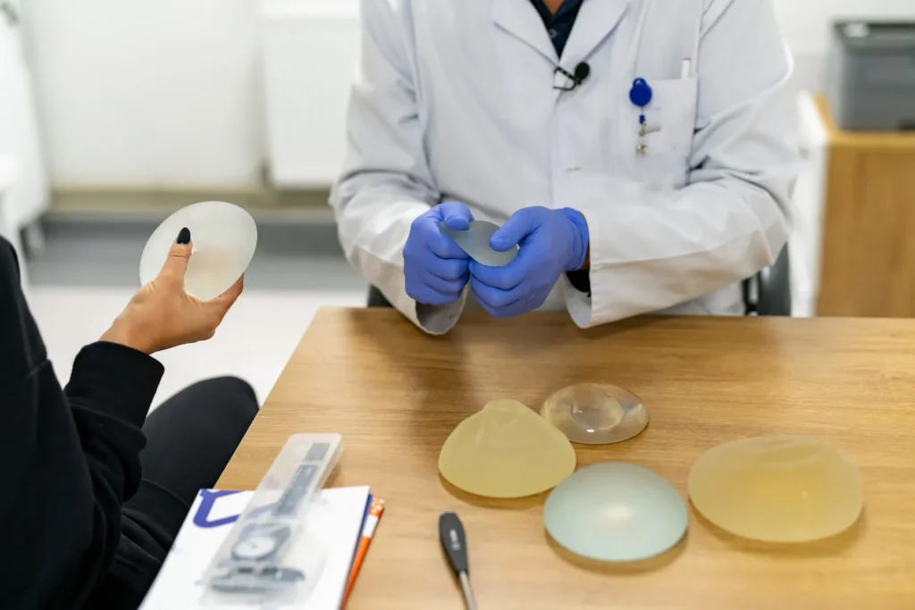 A doctor in a white coat holds a breast implant while discussing reconstruction options with a patient at a wooden desk.