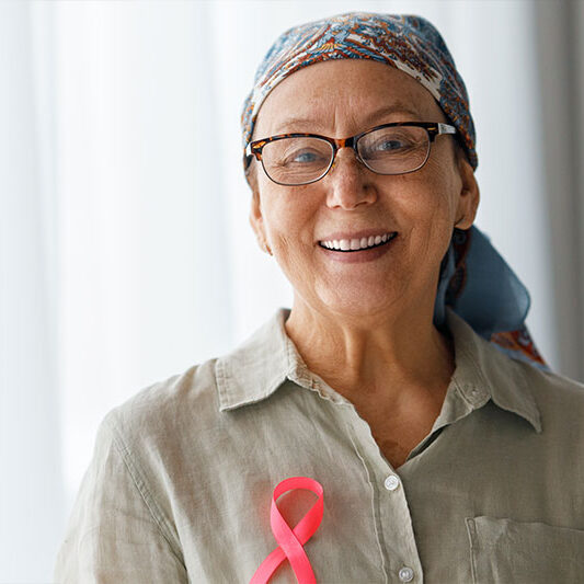 Smiling older woman with glasses and headscarf wears a pink ribbon on her shirt inside a Baton Rouge cancer hospital.