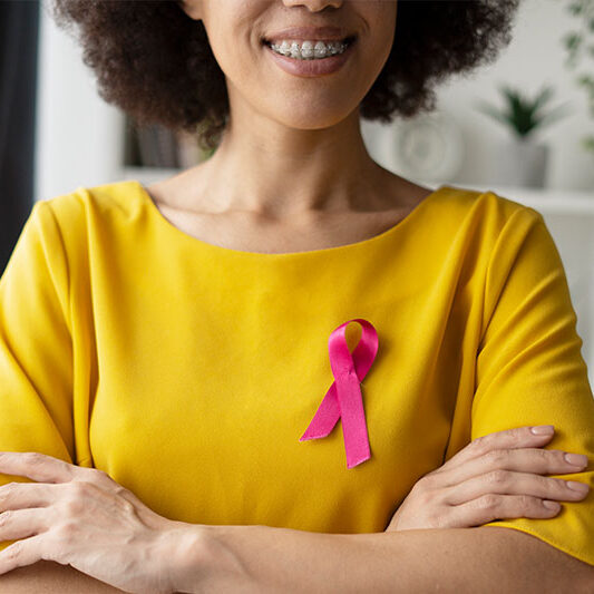 Woman in a yellow shirt with a pink ribbon stands with arms crossed, showing support at a recent breast cancer hospital.