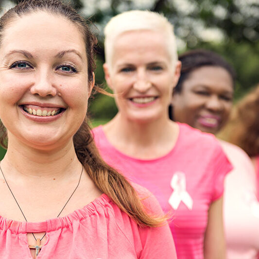 Four women in pink tops and ribbons smile together outdoors, showing support for a local breast cancer awareness event.