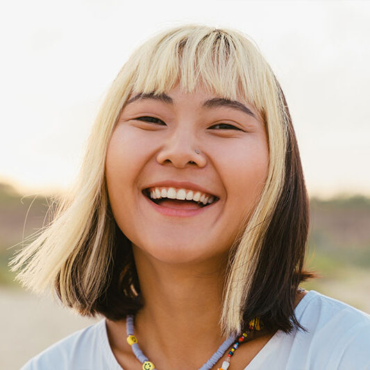 A person with two-toned hair smiles outside, dressed in a white top and beaded necklace near a hospital in Baton Rouge.