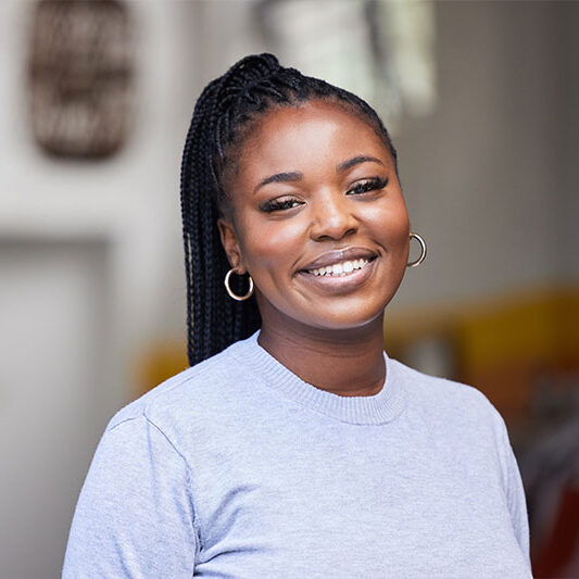 A smiling woman with braided hair, hoop earrings, and a light gray top stands in a Baton Rouge breast cancer hospital.