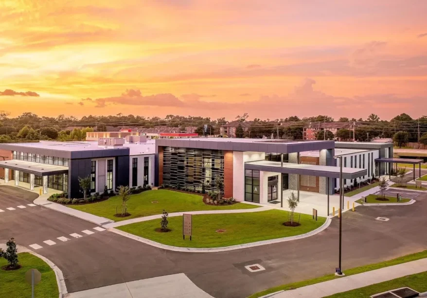 A single-story, modern hospital with large windows and landscaped grounds beneath a vibrant sunset sky.