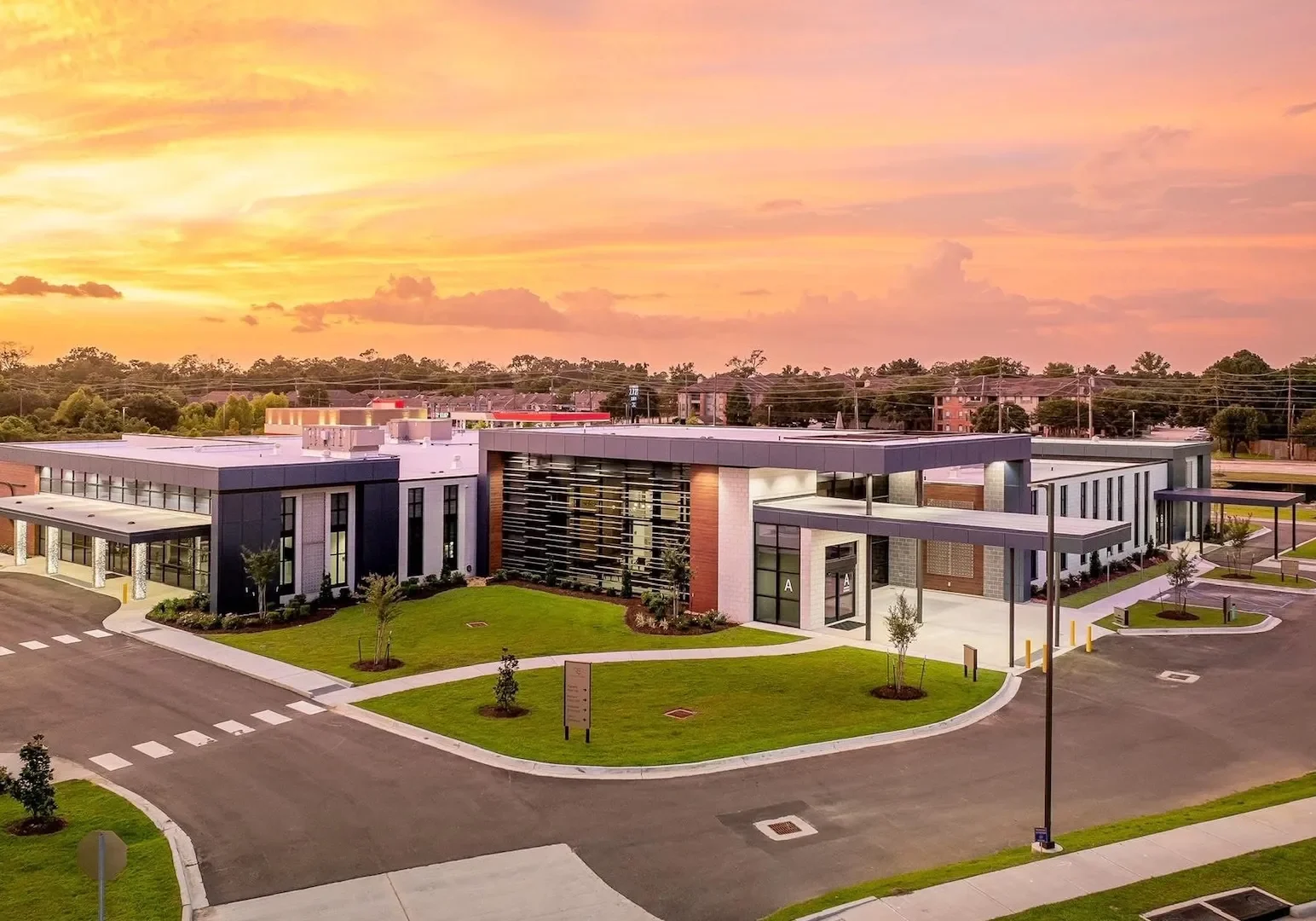 A single-story, modern hospital with large windows and landscaped grounds beneath a vibrant sunset sky.