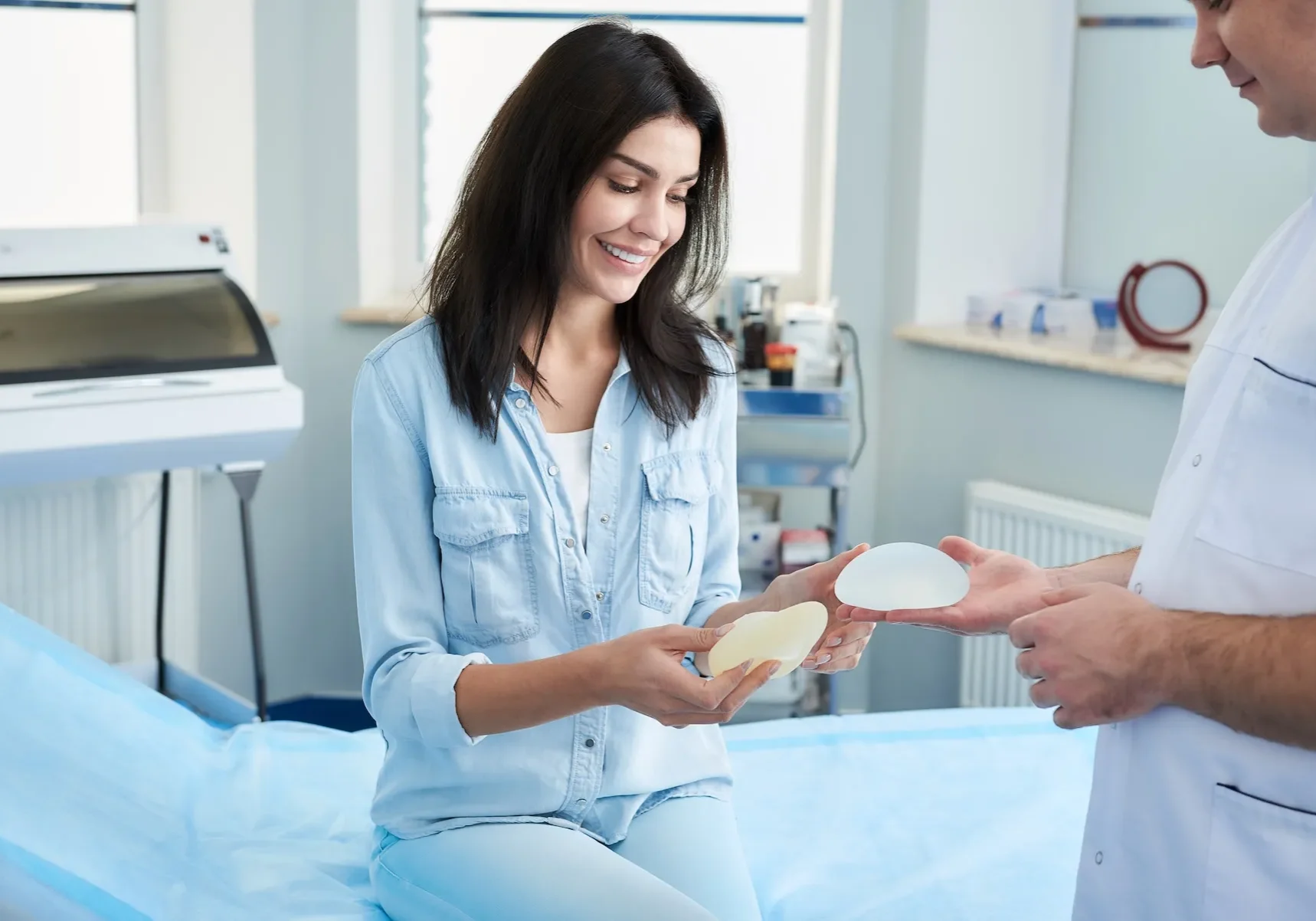 A woman smiles on an exam table, holding implants as a doctor displays additional samples beside her.