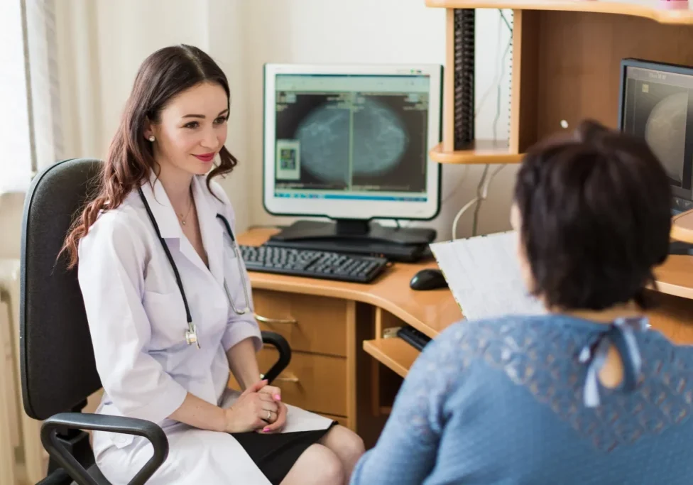 A doctor in a white coat talks with a patient, with medical images visible on computer screens in the background.