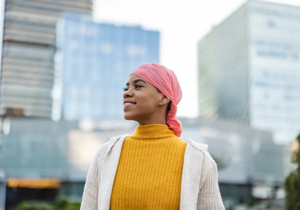 A woman in a pink headscarf and yellow sweater stands in front of tall modern buildings, every detail carefully captured.
