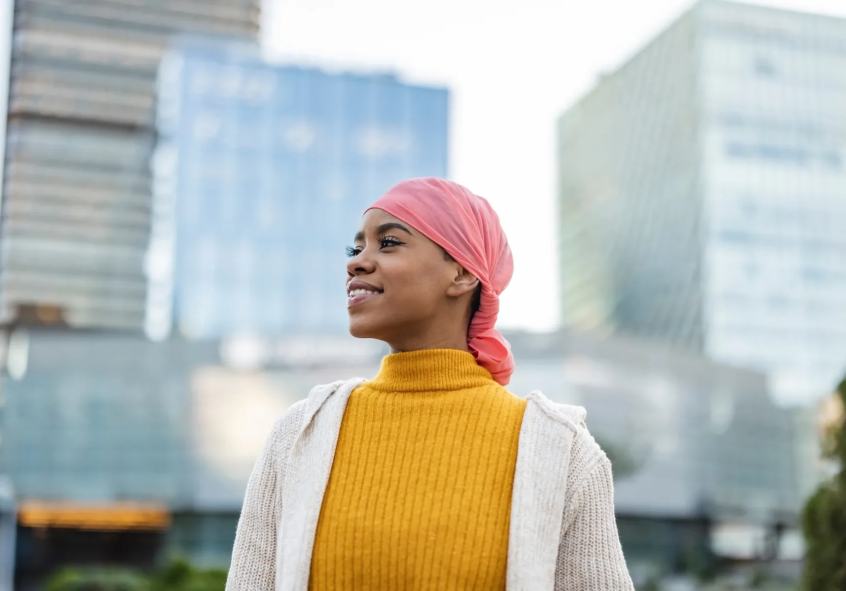 A woman in a pink headscarf and yellow sweater stands in front of tall modern buildings, every detail carefully captured.
