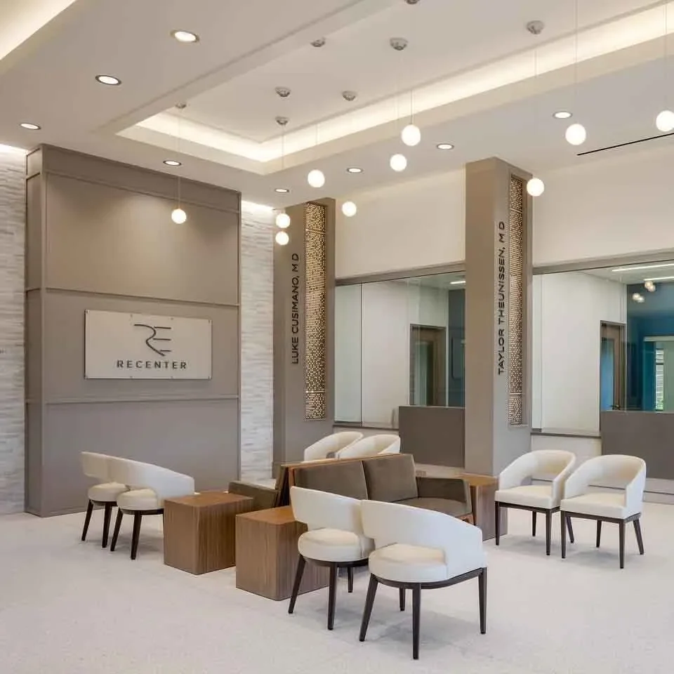Modern medical office waiting area at ReCenter featuring neutral-toned seating, wood accent tables, pendant lighting, and branded wall signage for physicians Luke Cusimano, MD and Taylor Theunissen, MD.