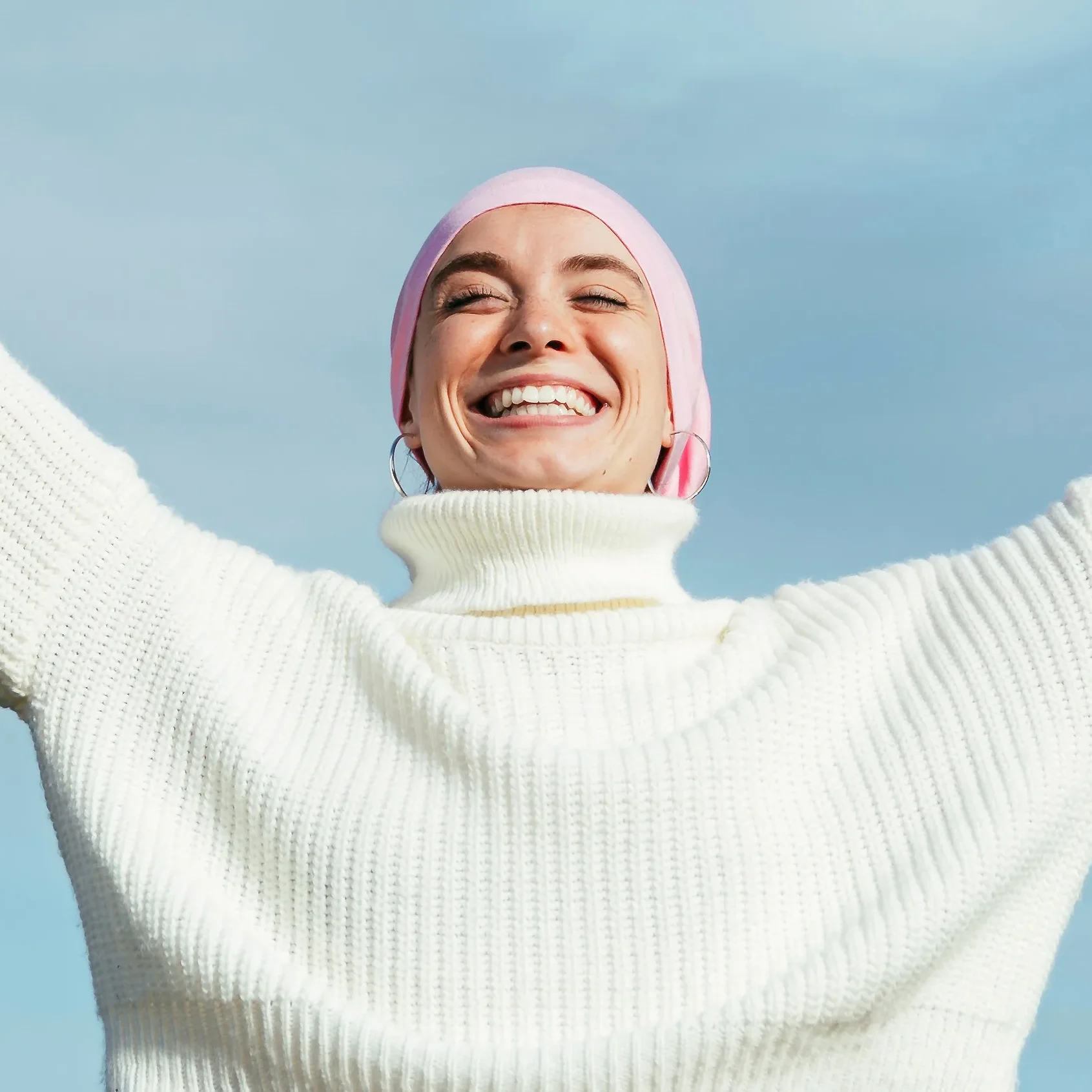 A woman in a white turtleneck and pink headscarf smiles with arms raised, celebrating under a clear blue sky.