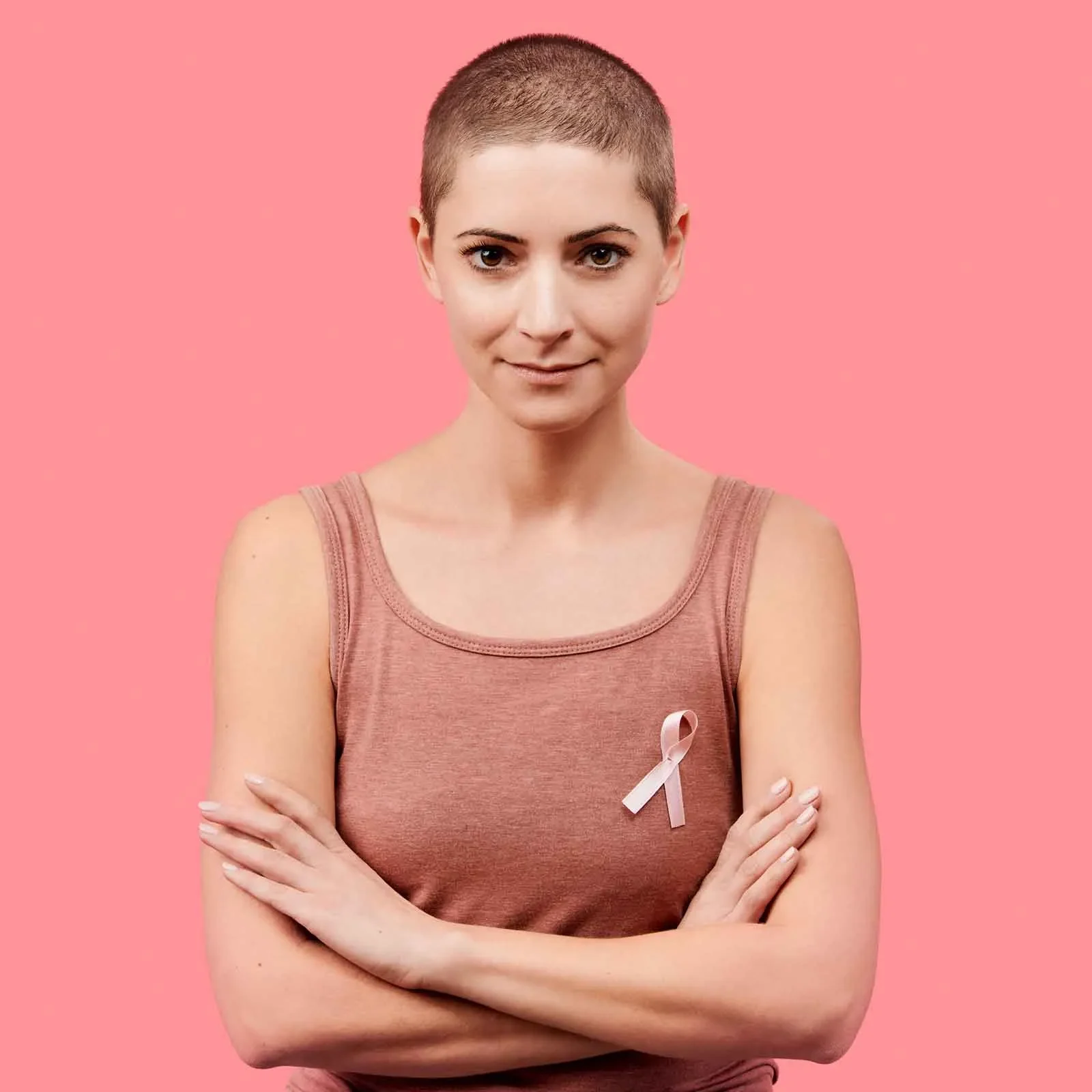 Woman with a shaved head wearing a pink ribbon and standing with arms crossed against a pink background, symbolizing breast cancer awareness and survivorship.