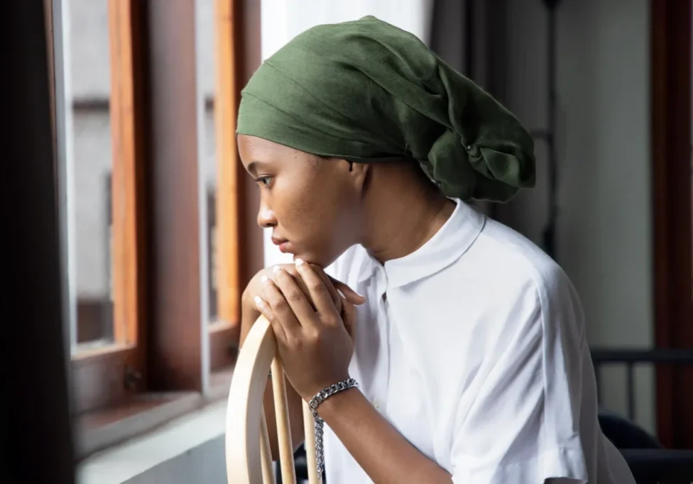 A woman in a white shirt and green headscarf sits indoors, looking out window.