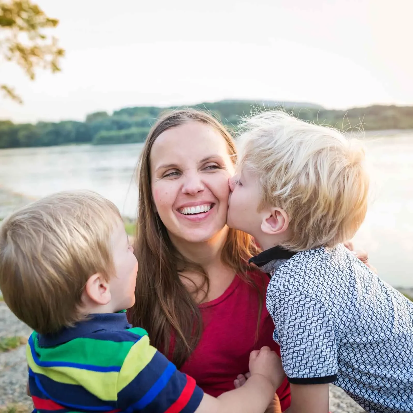 A smiling woman sits outdoors by the water while two young boys hug and kiss her on the cheek during a joyful family moment at sunset after receiving care at a Baton Rouge hospital.